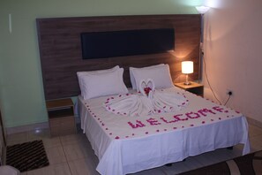 A neatly made bed with a welcome mat and heart-shaped towel arrangement, a bedside lamp, and a nightstand. - Football Hotel (Old-Yundum)