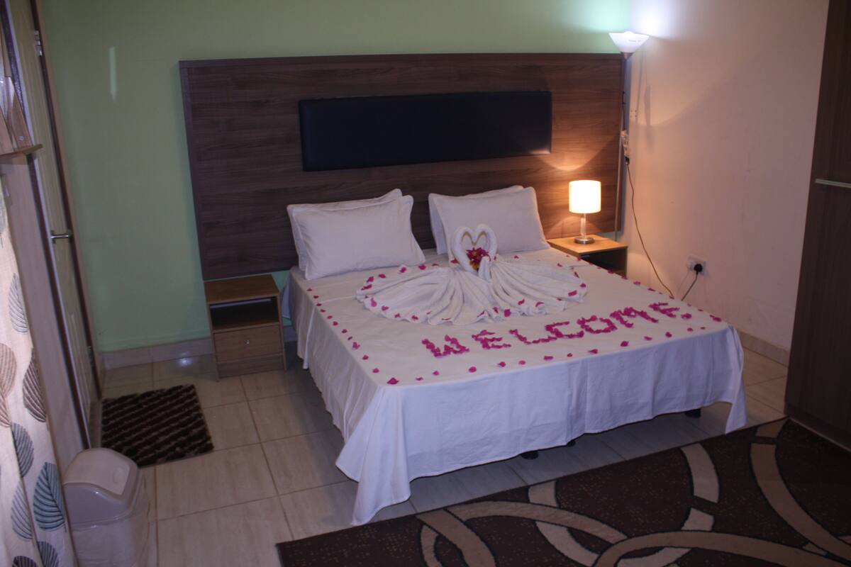 A hotel room with a bed, a bedside table with a lamp, and a welcome mat on the floor.