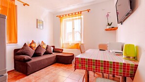 Un salon chaleureux avec un canapé marron, une table à manger recouverte d’une nappe à carreaux et un téléviseur fixé au mur.