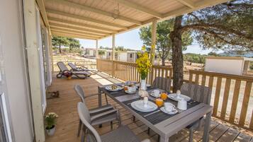 Un área de comedor al aire libre techada con una mesa preparada para el desayuno, rodeada de sillas, y vistas panorámicas a un lago y casas móviles.