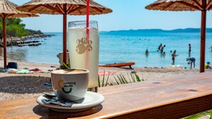A beachside café with a wooden table, a cup of coffee, and a glass of a beverage with a straw. Beach umbrellas and people in the water are visible in the background. - Mobile Homes at Camping Ugljan Resort (Preko)