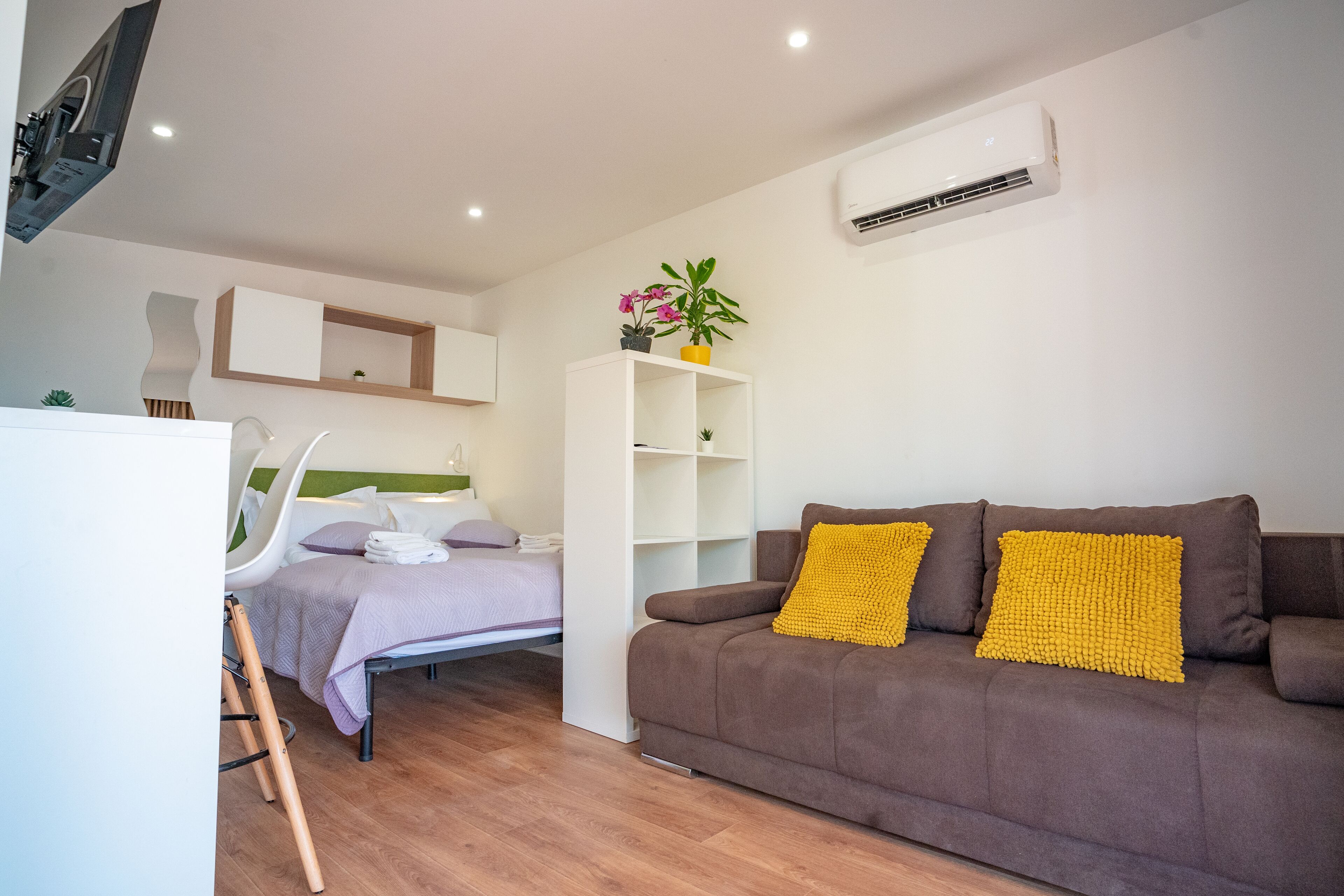 Una habitación moderna con un sofá marrón, una mesita de noche blanca, una cama con colcha morada, un armario blanco y una planta pequeña sobre un estante blanco.