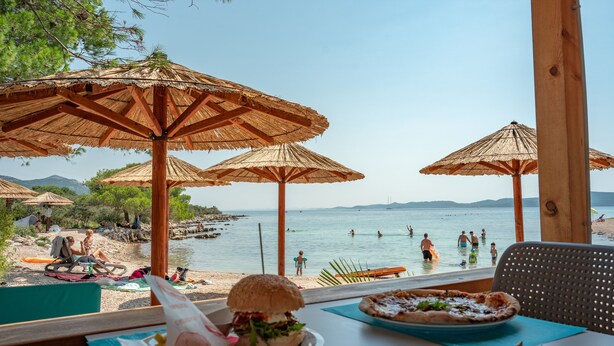 A beachside restaurant with outdoor seating, umbrellas, and a view of the sea. - Mobile Homes at Camping Ugljan Resort (Preko)