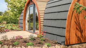 A modern, curved building with wooden and grey exterior walls, a large glass door, and a stone border around the garden area. - Chapel Pod (Saltash)