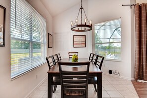 A dining area with a table, chairs, a chandelier, and large windows with blinds.