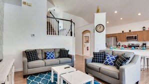 Ein modernes Wohnzimmer mit zwei grauen Sofas, einem blauen Teppich mit Muster und einem weißen Couchtisch. Im Hintergrund ist eine Küche mit Holzschränken und Edelstahlgeräten zu sehen.