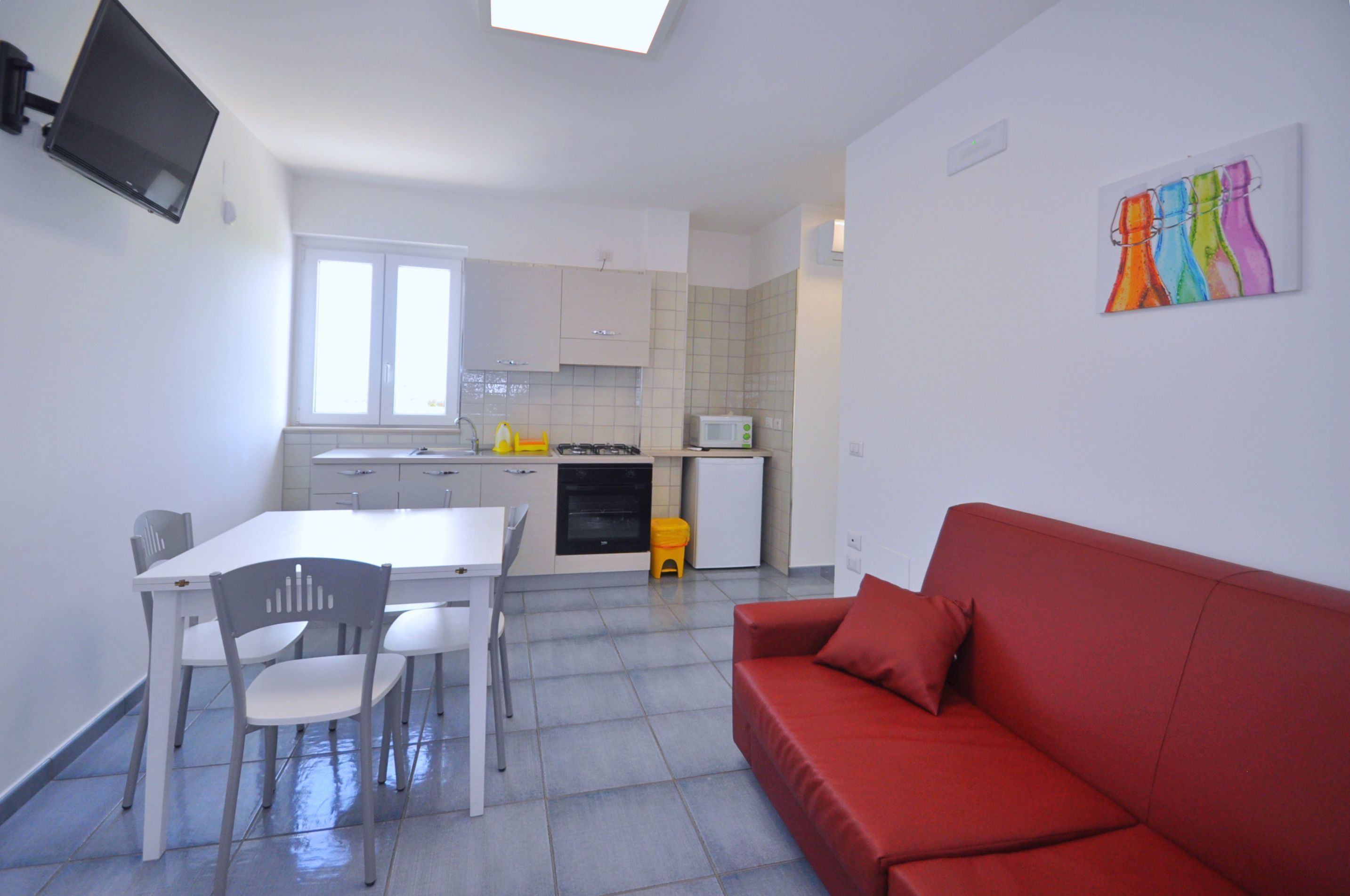 Un espace de vie compact comprenant un coin cuisine, un coin repas avec des chaises, un canapé rouge et une œuvre d’art colorée accrochée au mur.