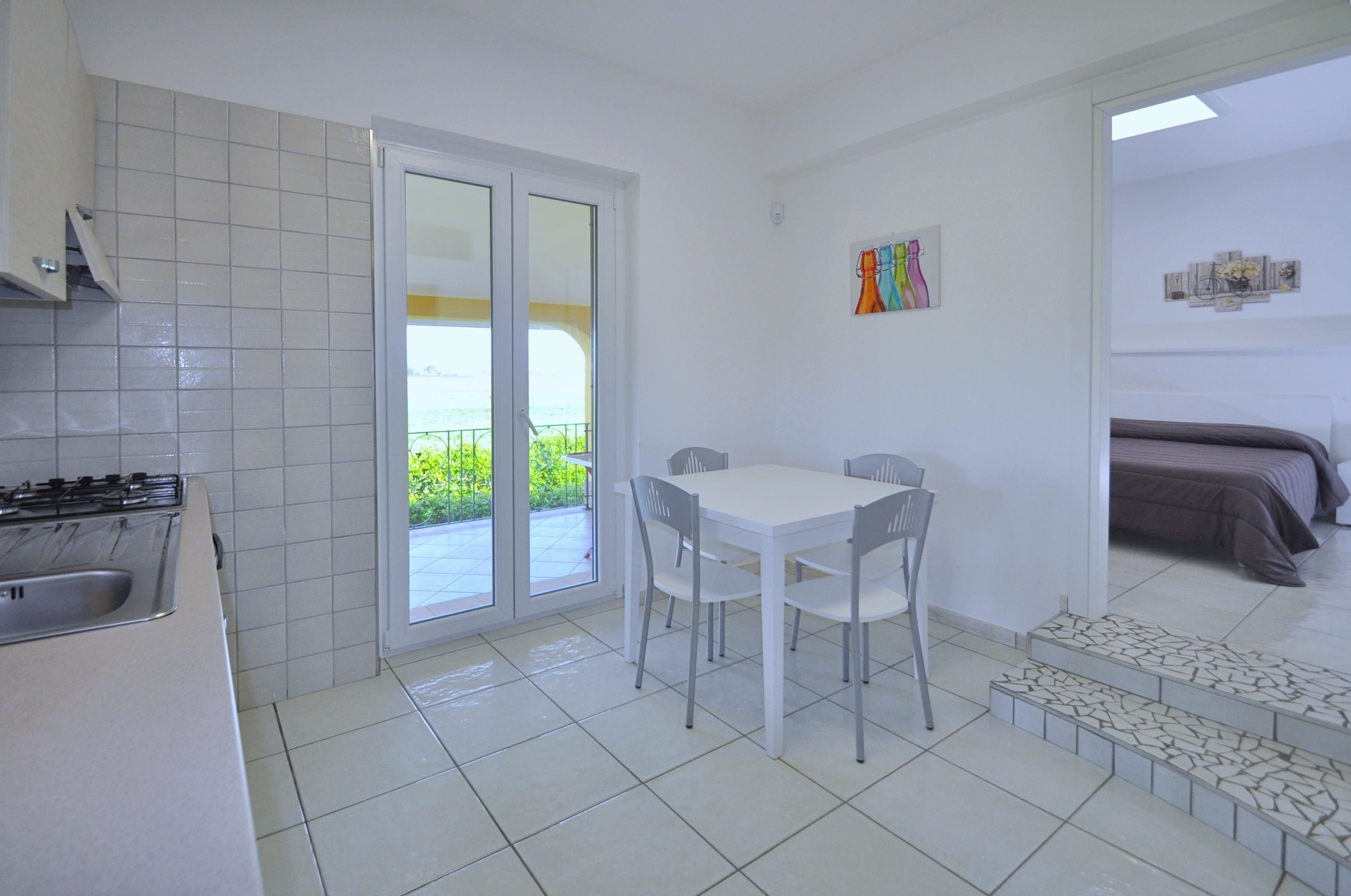 Une cuisine compacte équipée d’une cuisinière à gaz, d’un évier et d’un coin repas avec une table et des chaises. Une porte-fenêtre coulissante donne accès à un balcon offrant une vue sur la verdure.