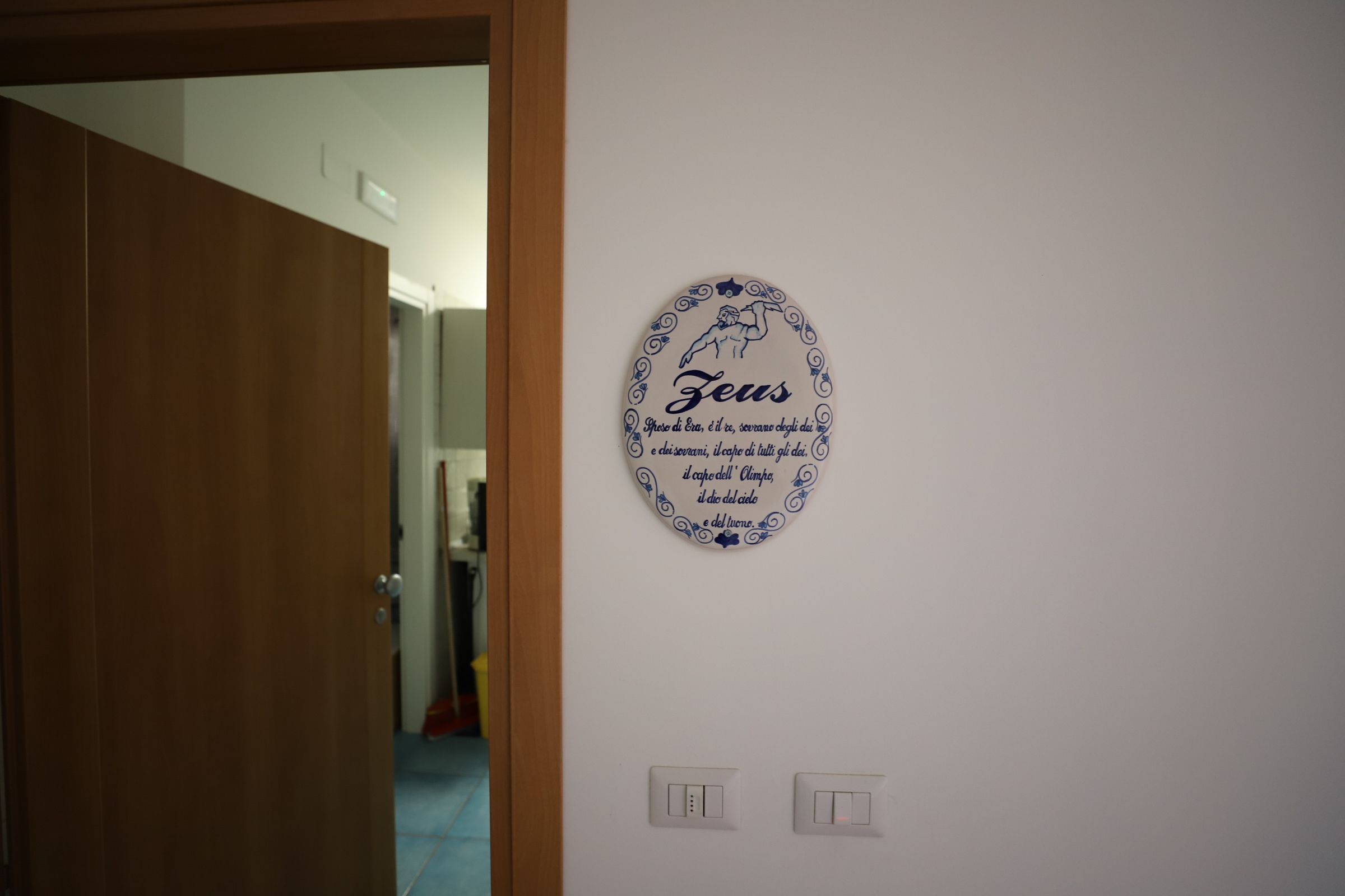 Pared con un plato decorativo que muestra la palabra 'Zeus' y texto italiano, y dos interruptores de luz debajo.