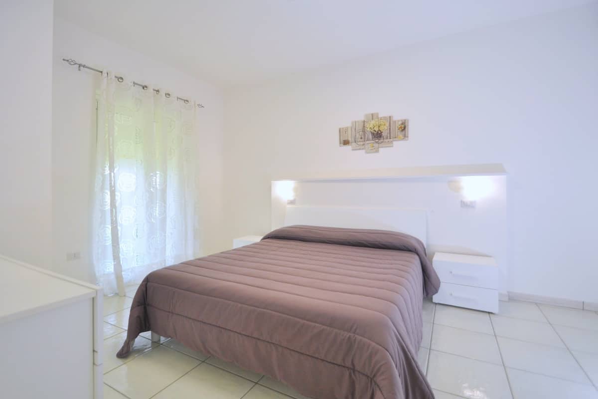 A bedroom with a bed, a nightstand, a wall-mounted light, and a window with sheer curtains.