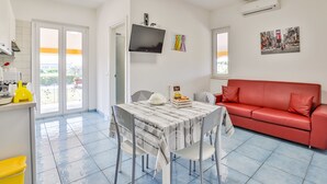 Un salon moderne avec un canapé rouge, une table à manger avec des chaises, un téléviseur fixé au mur et un balcon visible à travers la porte.