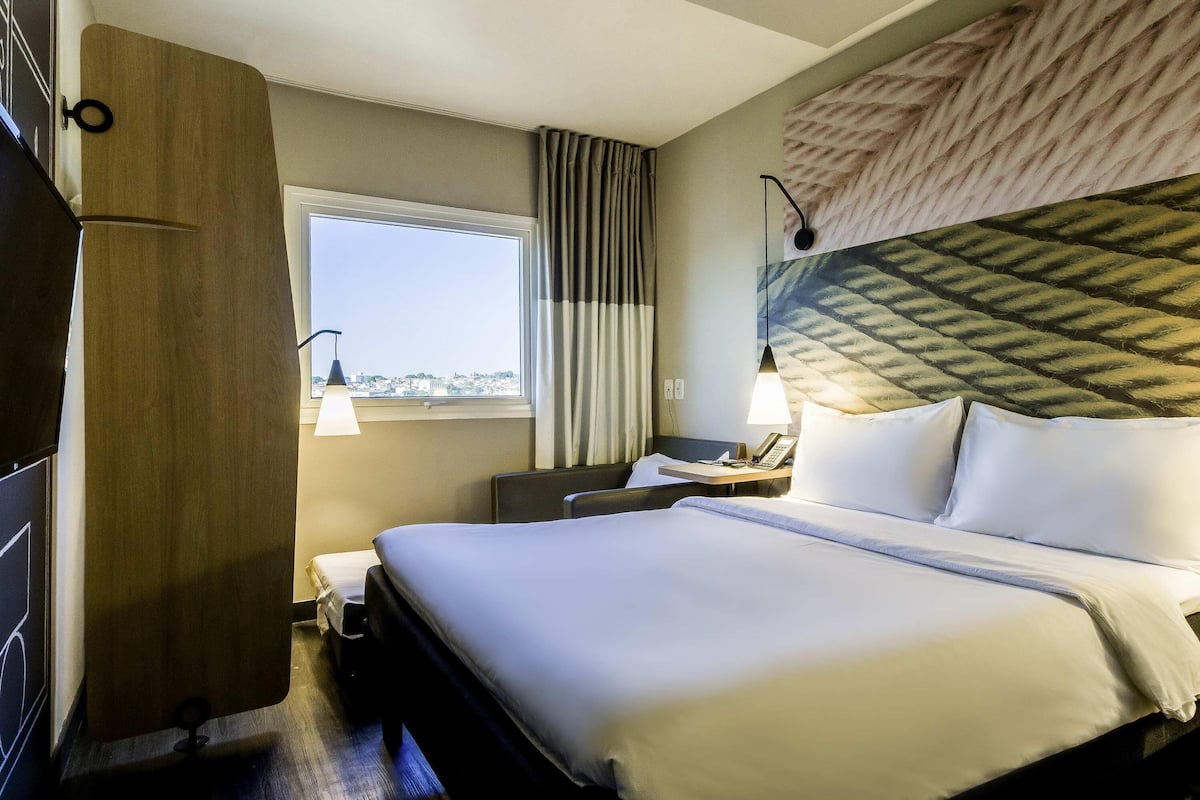 A hotel room with a large bed, a desk with a telephone, a window with curtains, and a wall-mounted lamp.