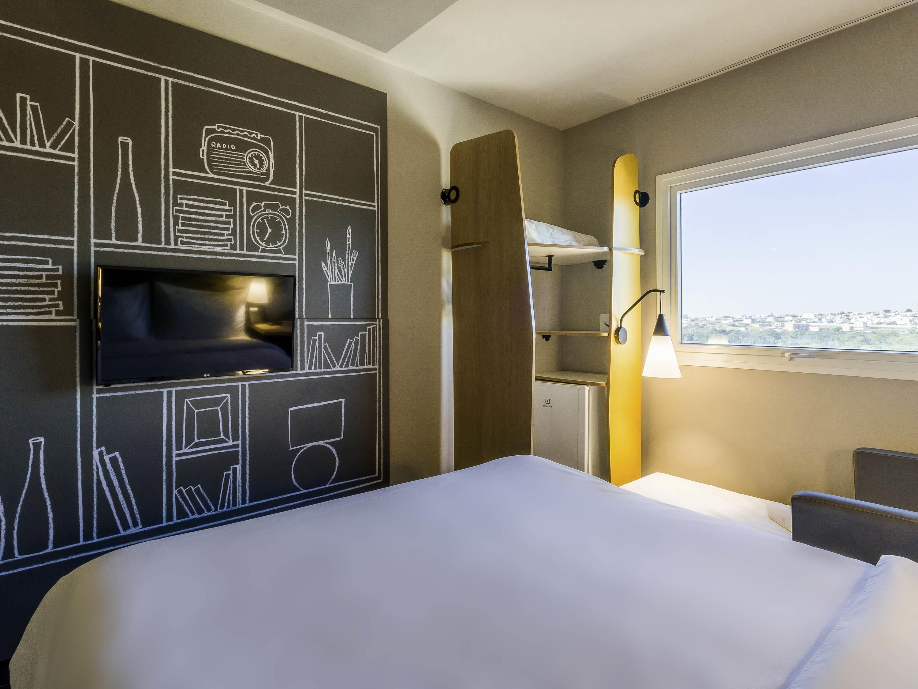 Una camera d'albergo con un letto, una TV, uno scaffale con dei libri, un'asse da surf e una finestra vista città.