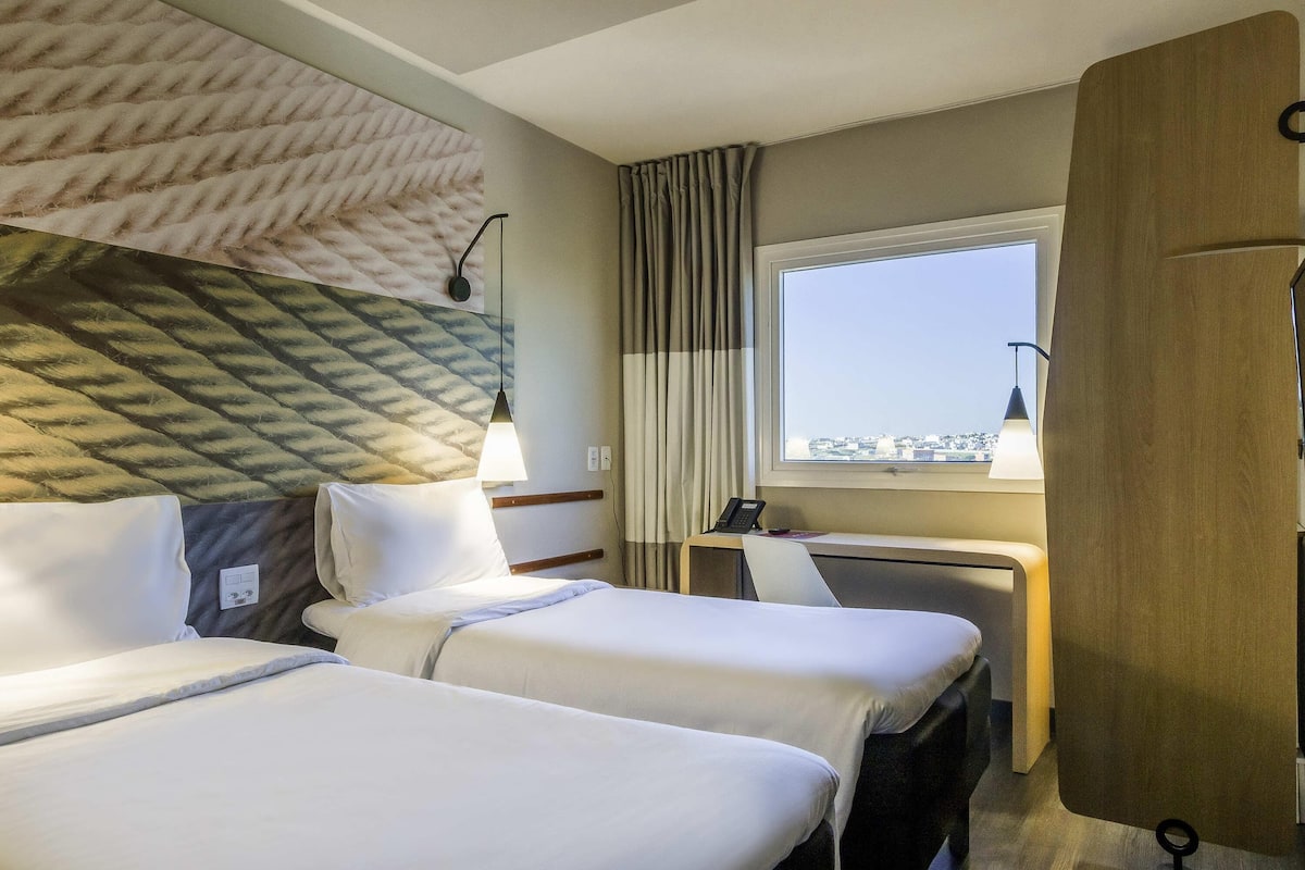 A hotel room with two beds, a desk, a window with a city view, and a headboard with a textured design.
