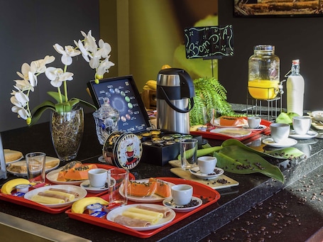 A breakfast buffet with various food items, coffee machines, and decorative elements like orchids and a neon sign.. ibis Americana