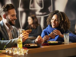 Dos personas cenando en un restaurante, con una mesa, una bebida y un plato de comida.