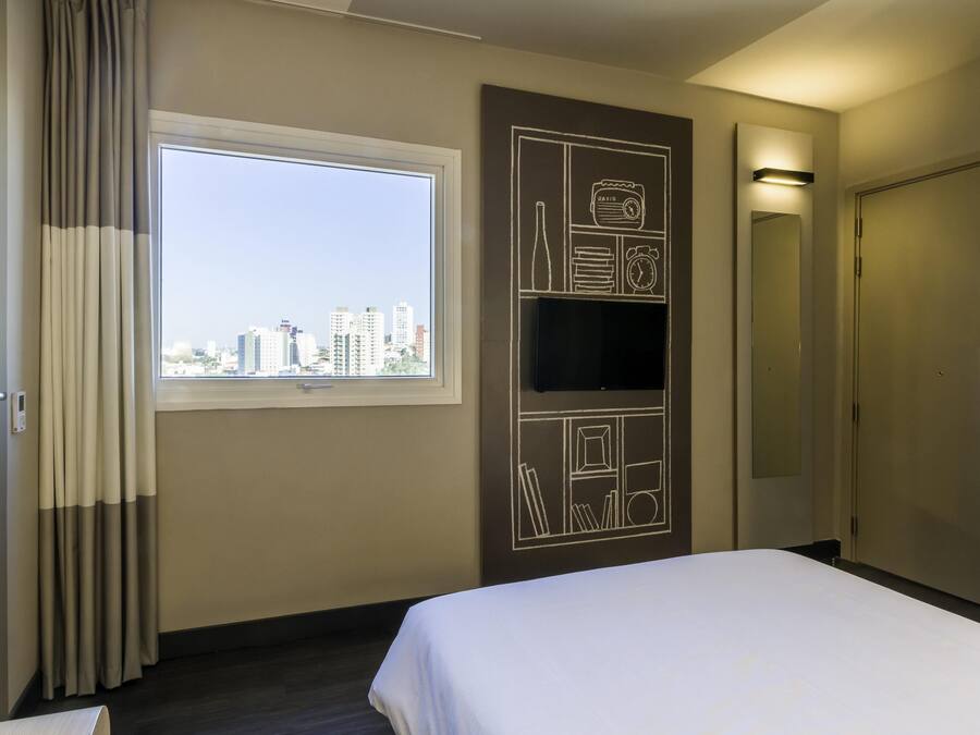 A hotel room with a large window, a flat-screen TV mounted on the wall, a bed, and a door.
