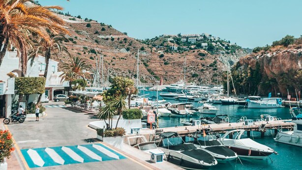 A marina with boats docked, a pedestrian path, and buildings on a hillside. - APARTAMENTOS PUERTO MARINA DEL ESTE (Almunecar)