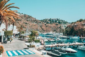 A marina with boats docked, a pedestrian path, and buildings on a hillside. - APARTAMENTOS PUERTO MARINA DEL ESTE (Almunecar)