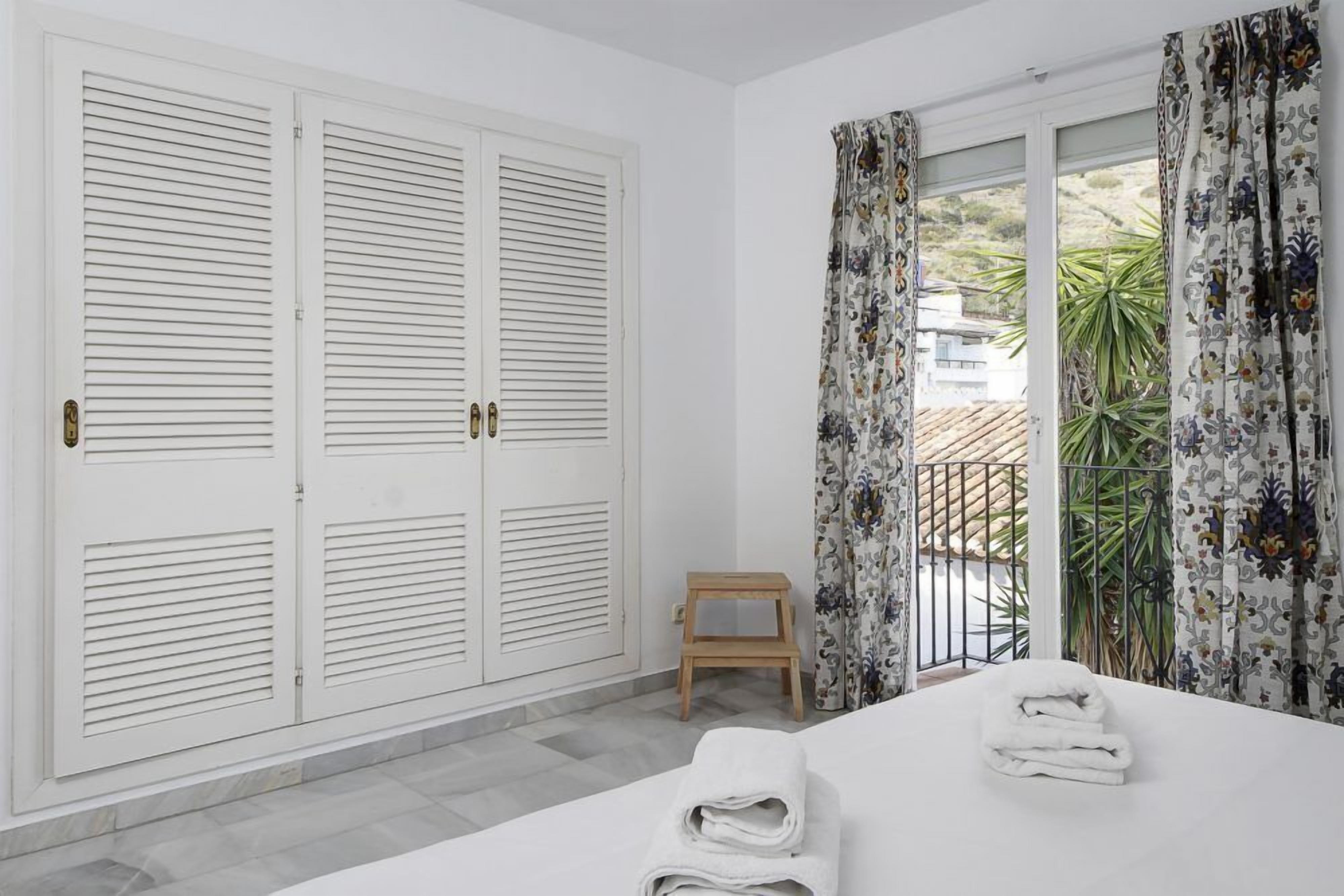 Habitación de hotel con cama, cortinas blancas, un taburete de madera y vistas al exterior.
