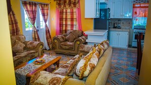 Un salon comprenant un canapé, des fauteuils et une table basse. Il y a également un coin cuisine avec des placards et une cuisinière.