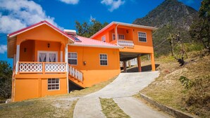 Une maison à deux étages, avec un toit rouge et des balustrades blanches, située sur une colline, avec une montagne en arrière-plan.