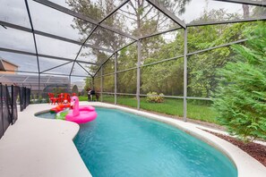 Una piscina con cerramiento de vidrio, rodeada de vegetación y un patio con sillas rojas.