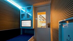 Una habitación con una cama, un escritorio y una puerta con ventana. Se observa una pantalla sobre la cama que muestra iconos.