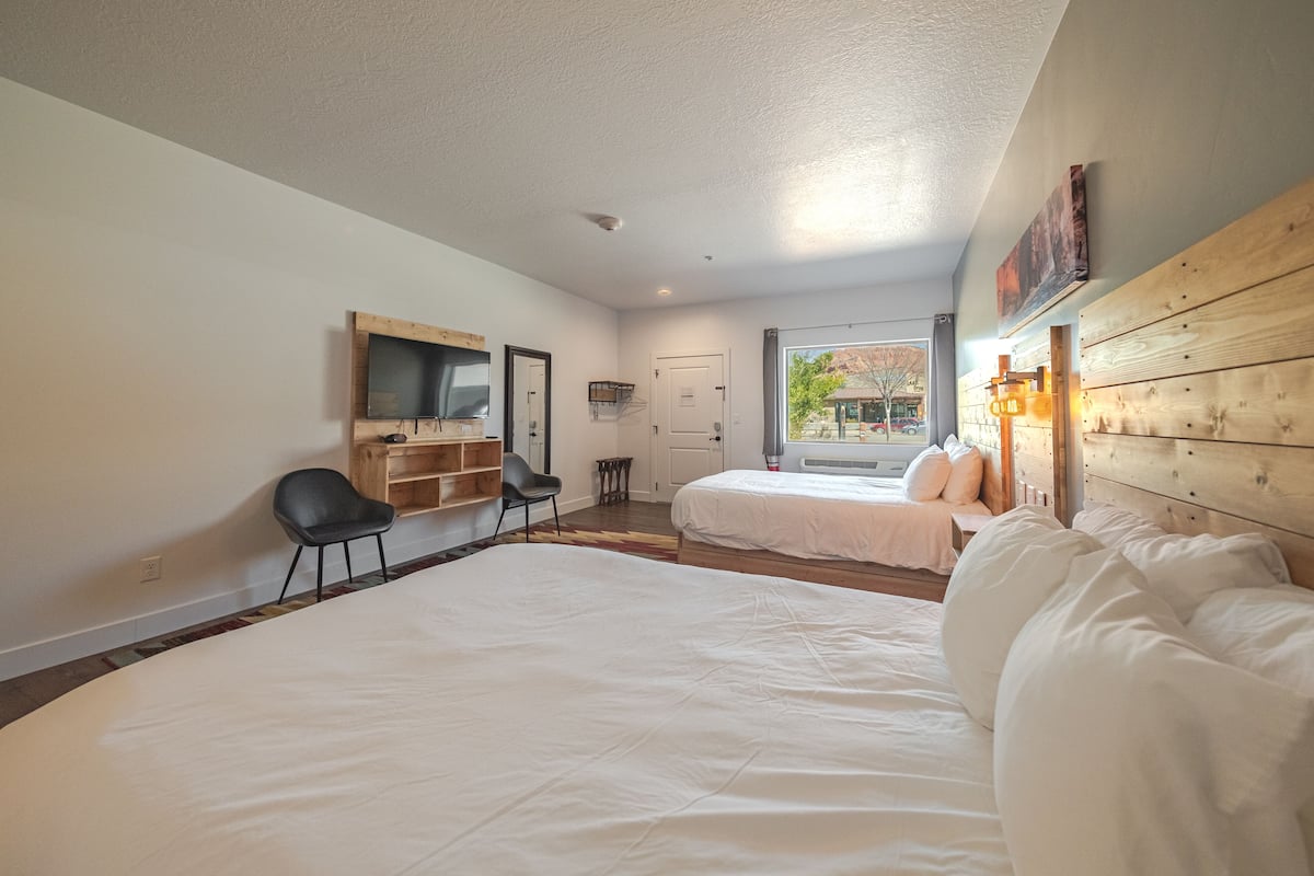A hotel room with two beds, a wooden headboard, a TV mounted on the wall, and a window with a view of the outside.