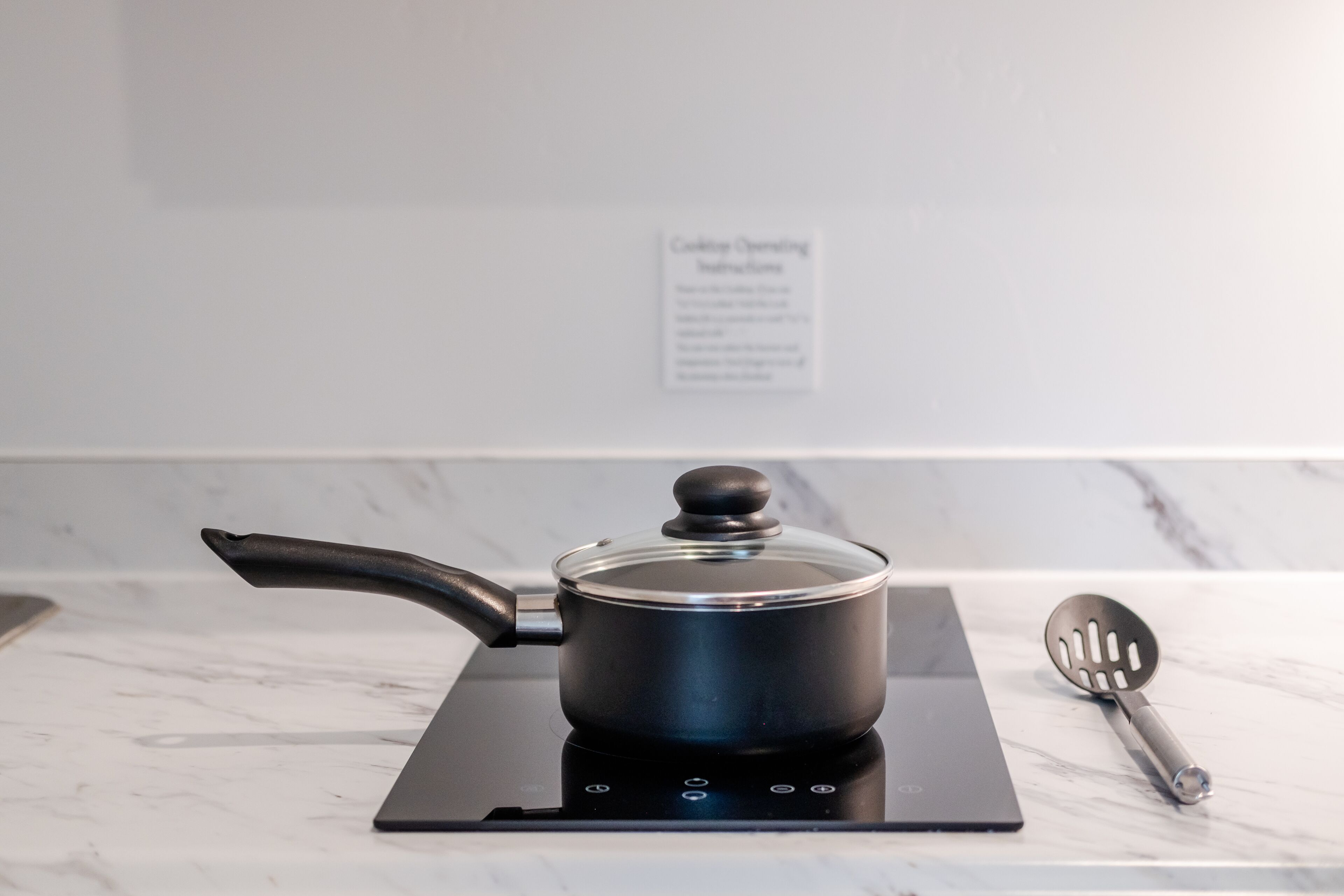 Een keuken met een moderne inductiekookplaat, een pan met deksel, en een spatel op een marmeren aanrecht.