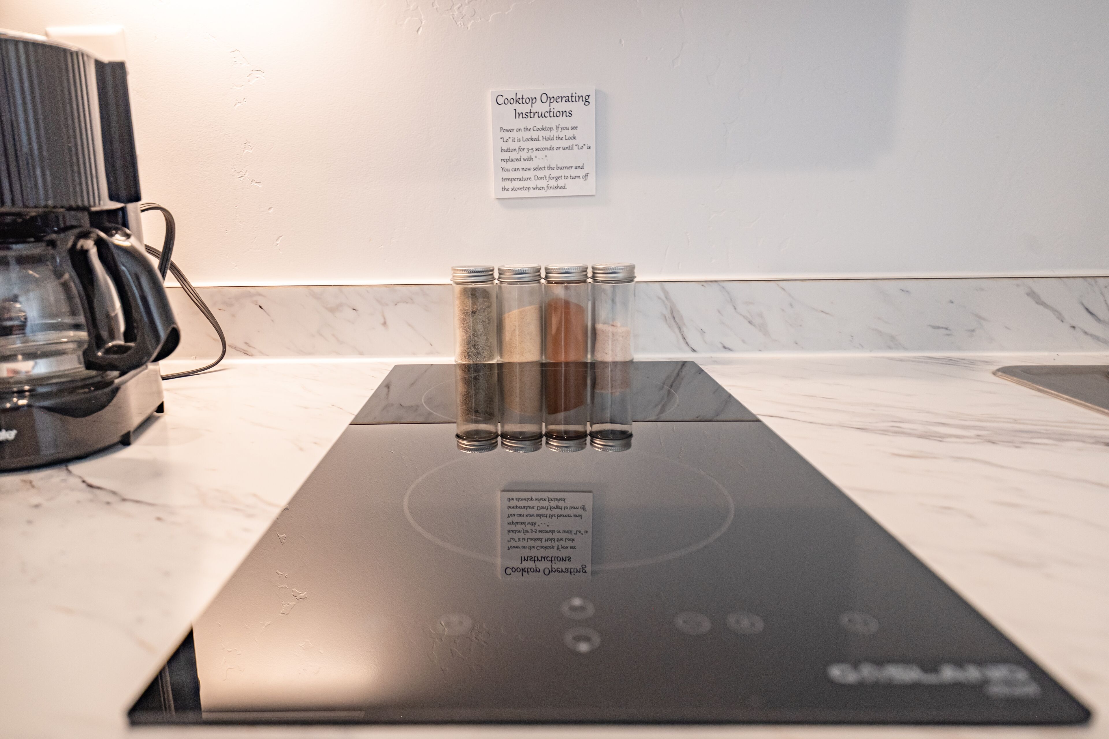Une plaque de cuisson avec quatre pots d’épices, une machine à café et un panneau explicatif fixé au mur.