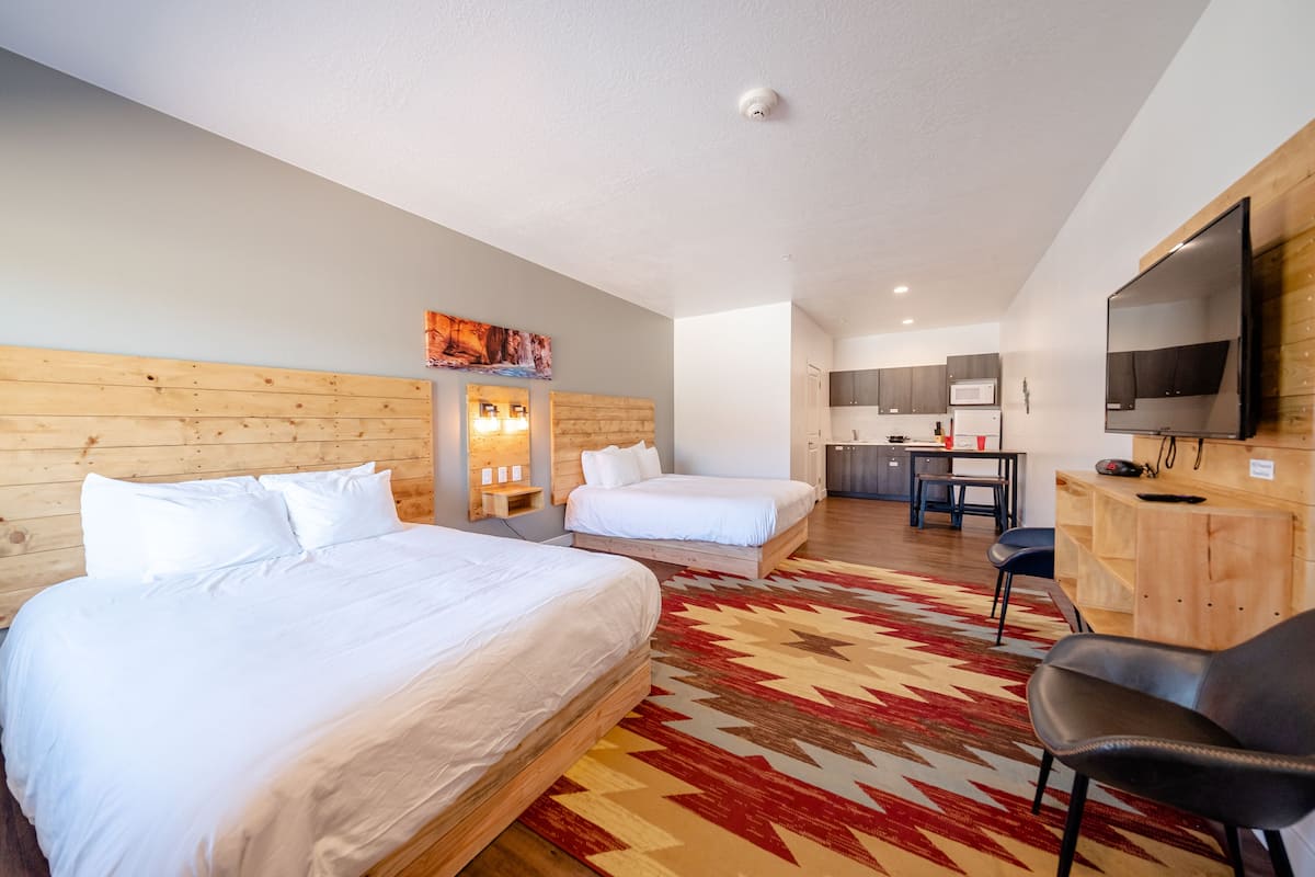 A hotel room with two beds, a desk, a TV, and a kitchen area.