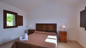 A bedroom with a wooden headboard, a bed with a brown bedspread, two bedside tables with lamps, and a window with a view of greenery. - Appartamenti in Gallura (Badesi)