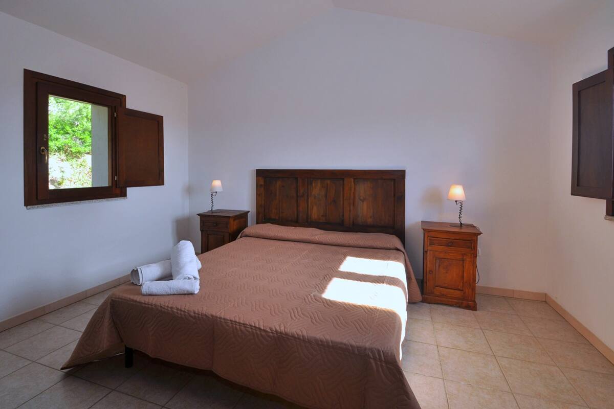 A bedroom with a wooden headboard, a bed with a brown bedspread, two bedside tables with lamps, and a window with a view of greenery.