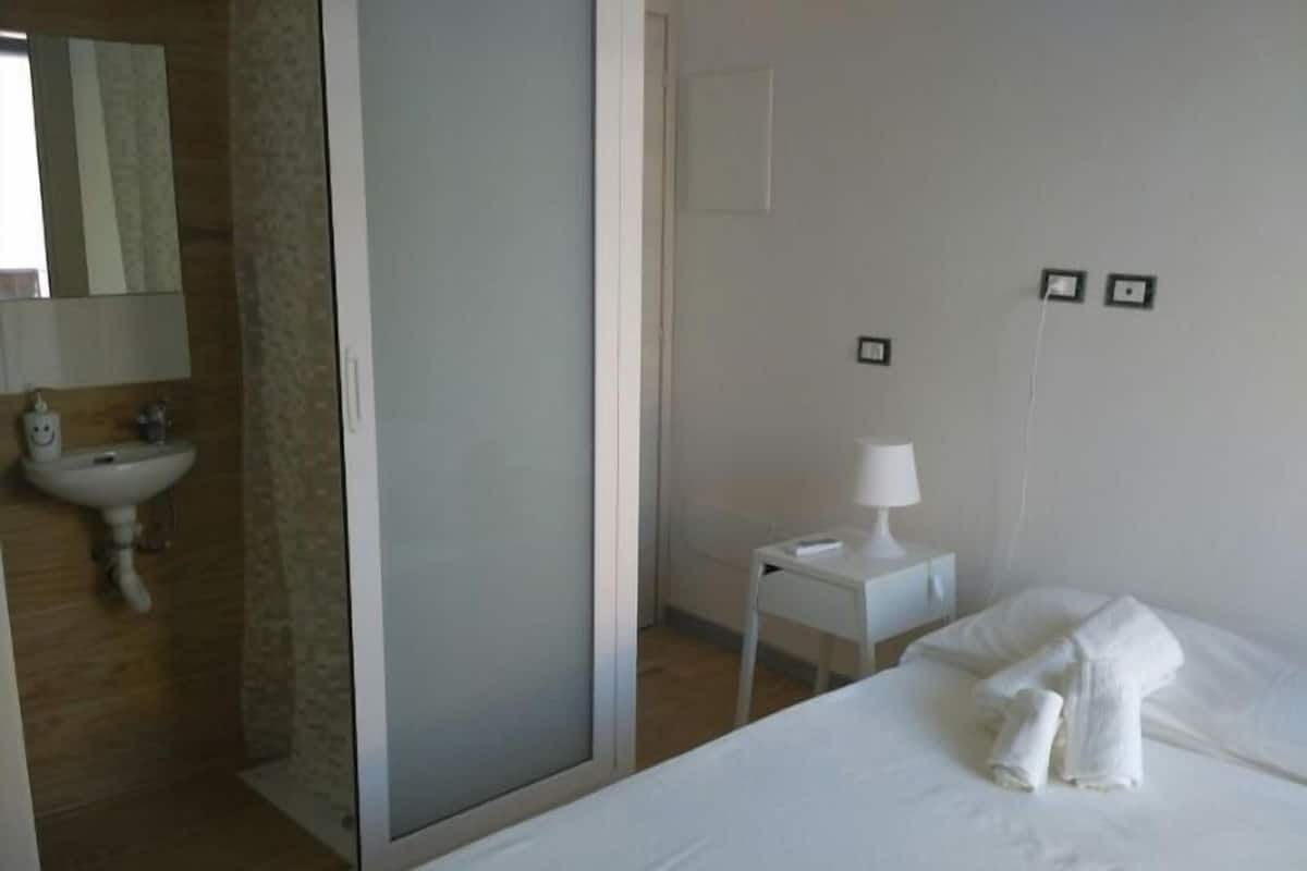 A hotel room with a bed, a bedside table with a lamp, a small white nightstand, a bathroom with a sink and mirror, and a sliding door.