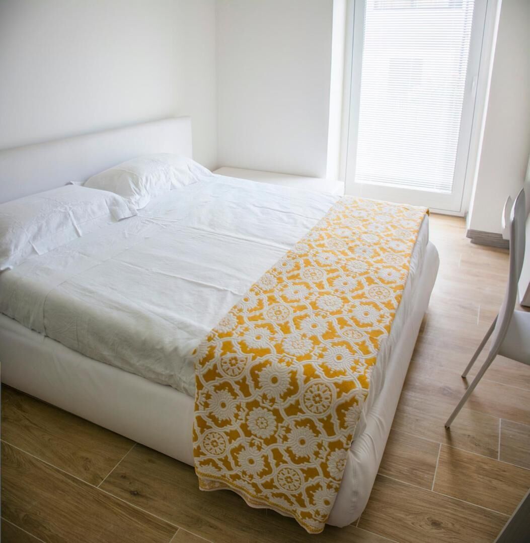 Un lit bien fait, recouvert d’une couvre-lit à motifs blancs et jaunes, dans une pièce lumineuse dotée d’un plancher en bois et d’une fenêtre avec des stores.