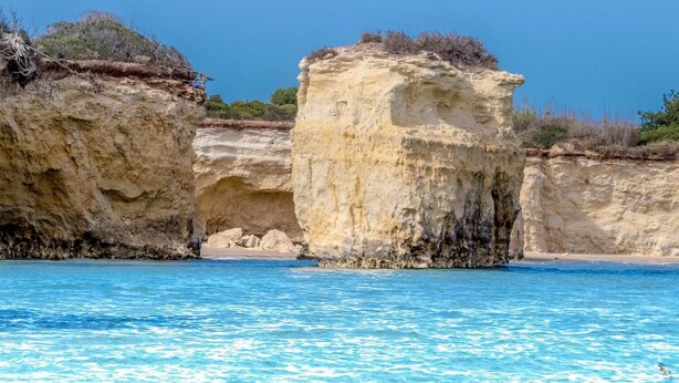 Escena costera con agua azul cristalina, una playa arenosa y acantilados rocosos.