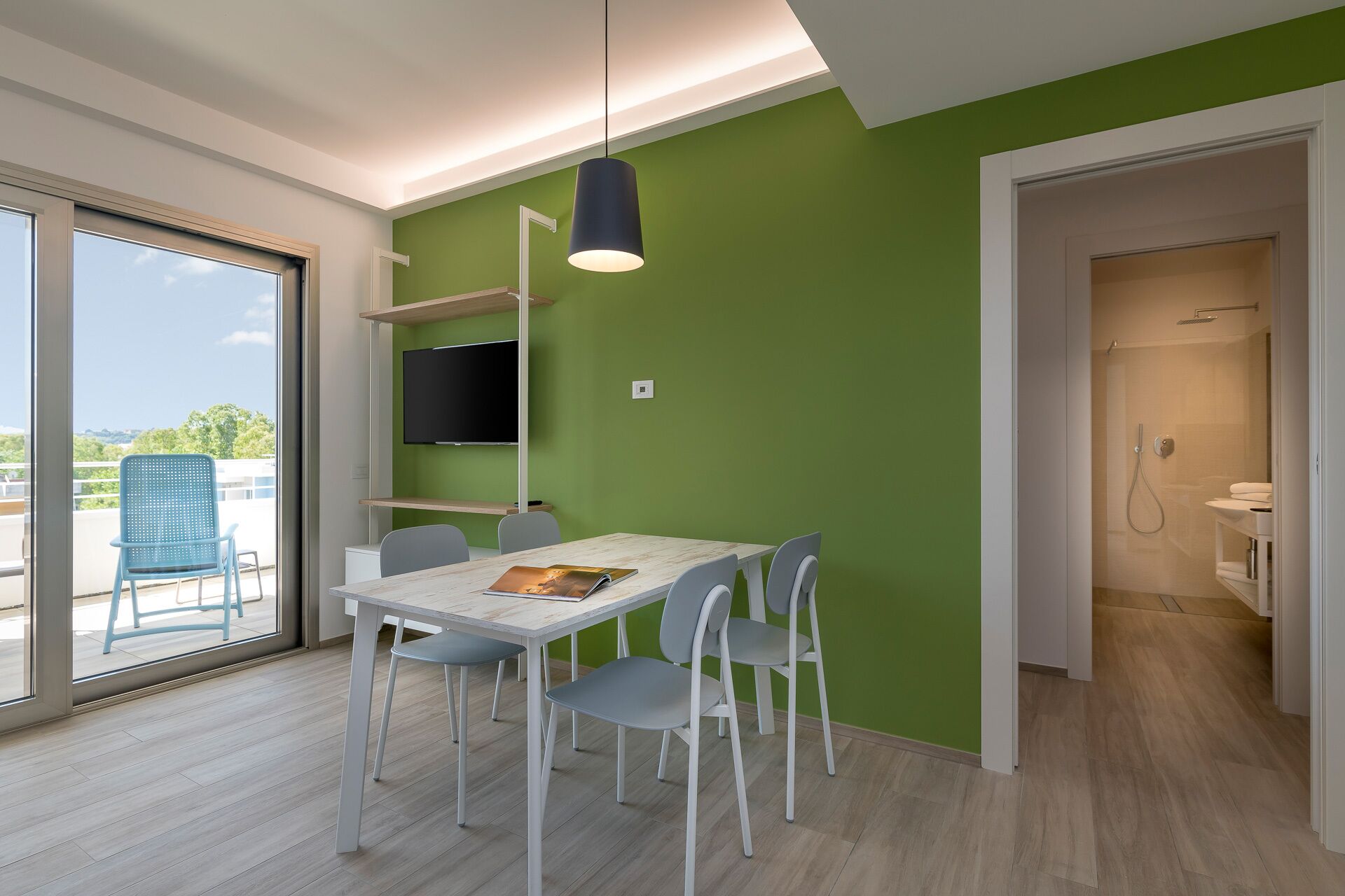 Quarto moderno com uma mesa e cadeiras brancas, televisão montada na parede e uma parede verde decorativa.