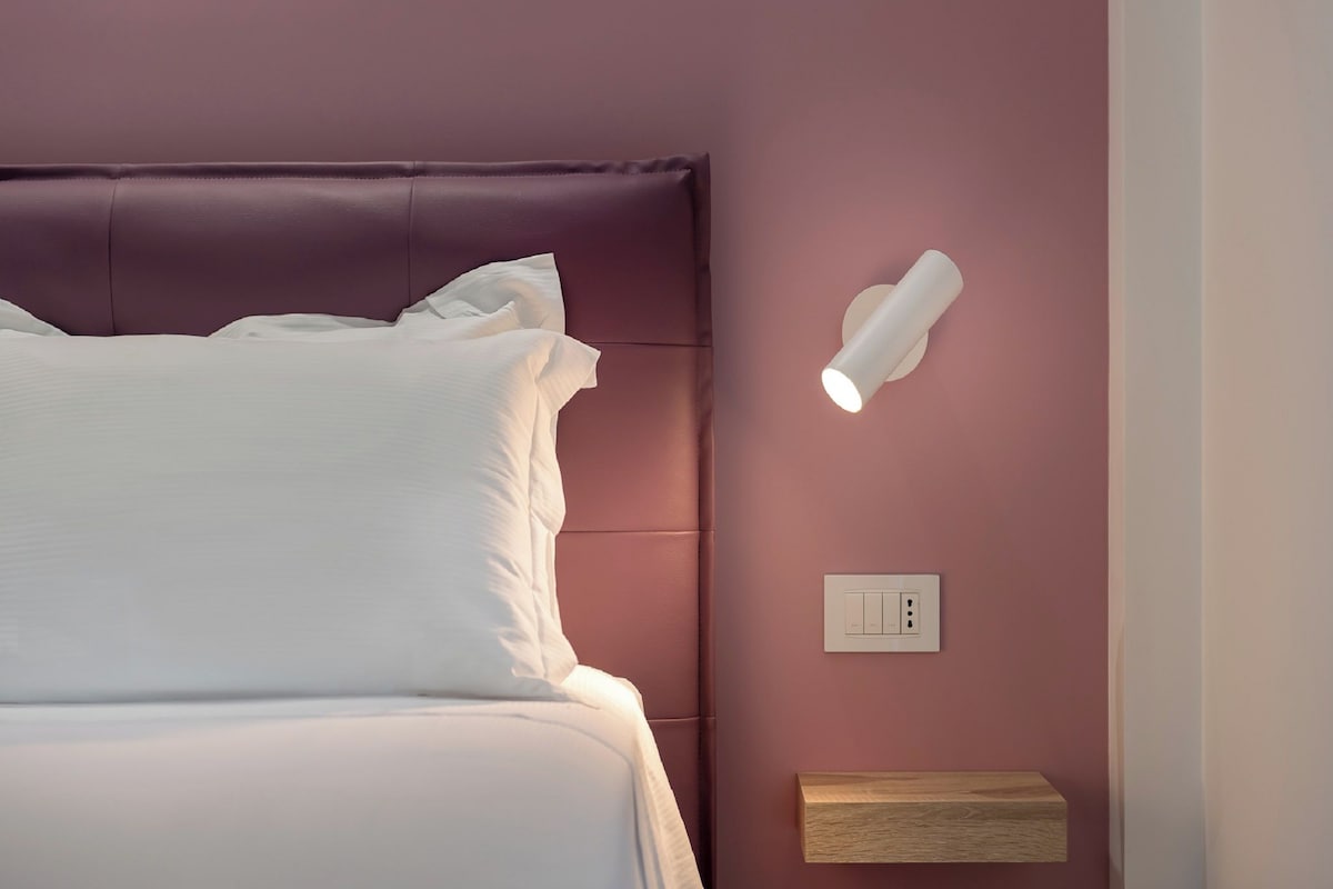 Una camera da letto con un letto, una lampada a parete, un interruttore e una mensola in legno.