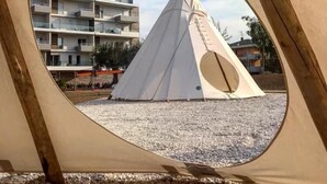 Vista a través de un marco de madera de un tipi blanco sobre grava, con un edificio de varios pisos al fondo.