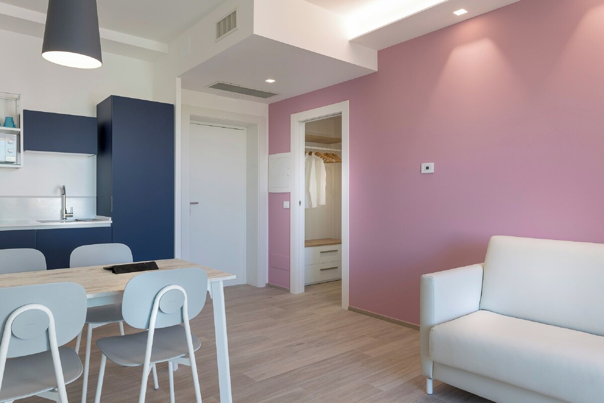 Een moderne kamer met een houten eettafel, witte stoelen, een witte bank en een roze accentmuur.