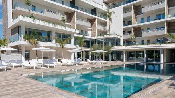 Un hotel moderno con zona de piscina en la azotea, que cuenta con tumbonas y sombrillas, rodeado de un edificio de varios pisos con balcones.