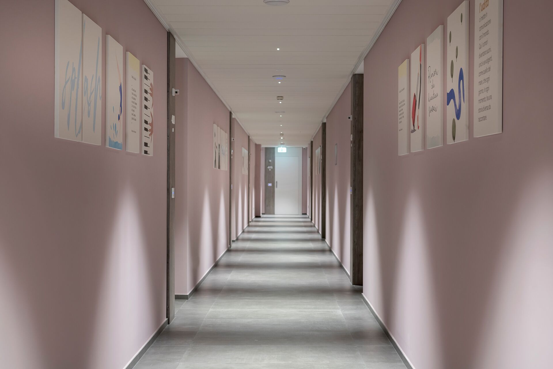 A long, narrow hallway with light pink walls and a grey floor, lined with framed artwork on both sides.
