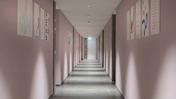 A long, narrow hallway with light pink walls and a grey floor, lined with framed artwork on both sides.