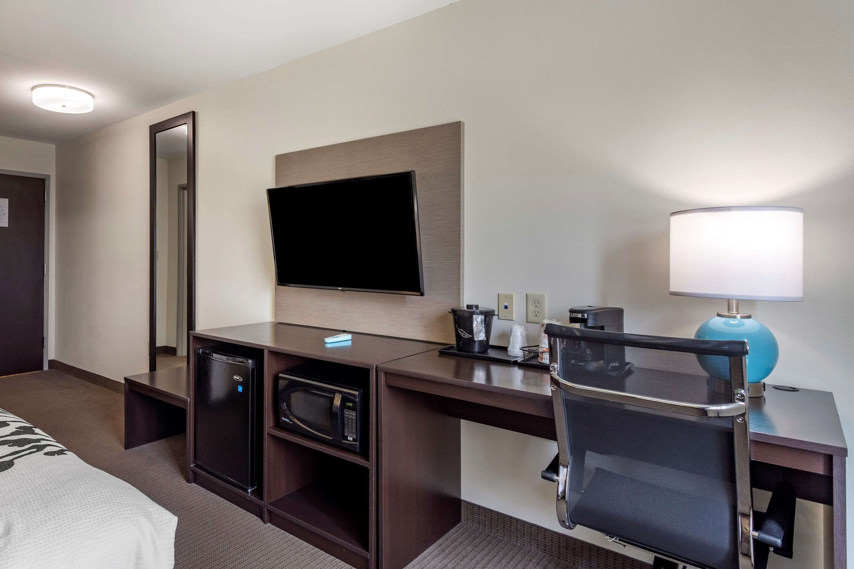 Una habitación de hotel con cama, escritorio con computadora, televisor, lámpara, microondas y refrigerador pequeño.