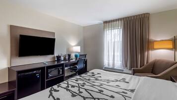 A hotel room with a bed, a desk with a flat-screen TV, a chair, a desk lamp, and a window with curtains.