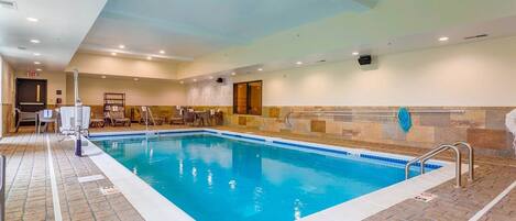 An indoor swimming pool with clear water, tiled walls, and a tiled floor. There are lounge chairs and a small table near the pool.