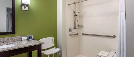 A bathroom with a green wall, a sink with a mirror, a white chair, a shower with a grab bar, and a white curtain.