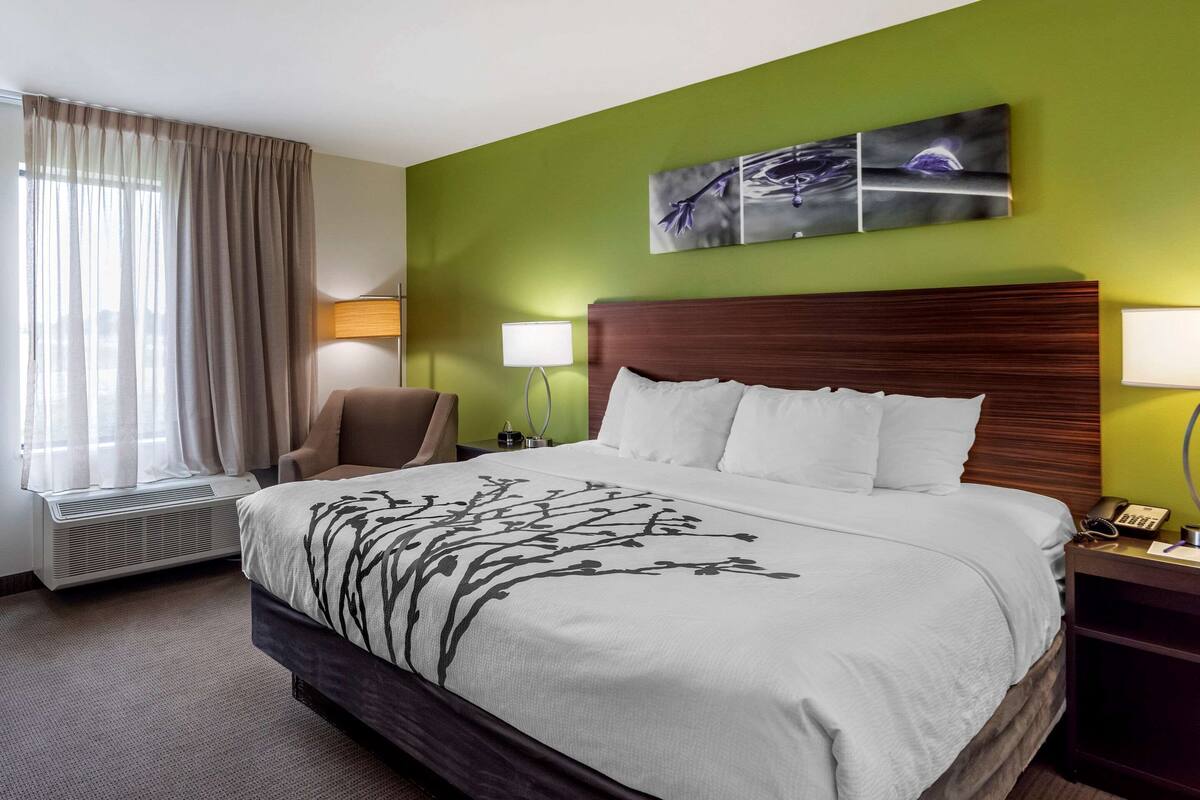 Quarto de hotel com uma cama grande, uma cadeira, uma mesa de cabeceira com abajur e uma janela com cortinas.