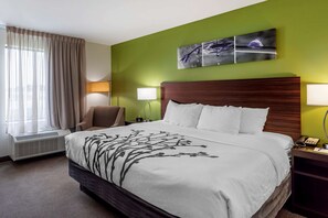 A hotel room with a large bed, a chair, a bedside table with a lamp, and a window with curtains.